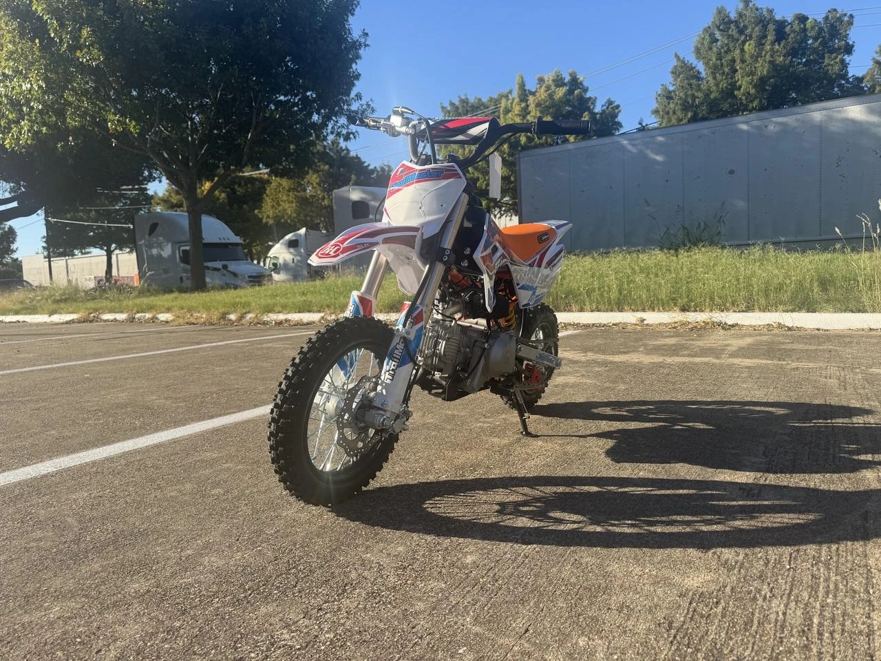 TrailMaster SK110S 110cc Semi-Automatic Kids Dirt Bike, off-road motorcycle with orange and white body design, equipped with knobby tires and front suspension, on a paved outdoor surface.