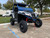 TM 4x4 SportsCross 1000 Side by Side, rugged off-road all-terrain vehicle with large tires, black and blue color scheme, reinforced roll cage, and front suspension in a parking lot with trees in the background.