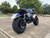 TM Hurricane 200X Upgraded Mini Bike, off-road mini motorcycle with large tires, blue and black body, and chrome accents, parked in an outdoor lot with trees in the background.