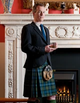 Man in blue and green tartan traditional kilt with jacket and sporran by a fireplace