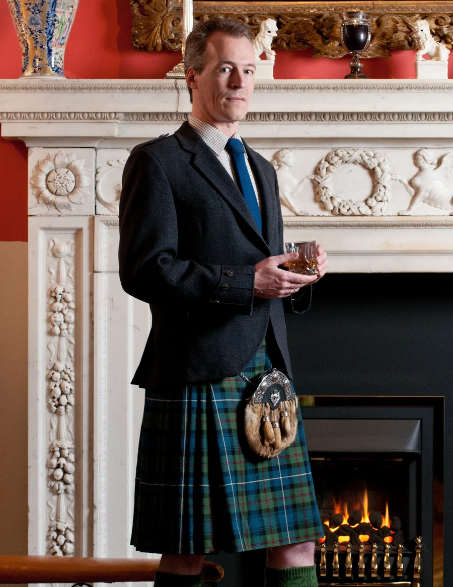 Man in blue and green tartan traditional kilt with jacket and sporran by a fireplace