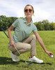 A light green polo shirt is worn by a woman crouching on grass, holding a golf ball, with sunglasses and casual trousers.