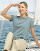 A woman wearing a light blue premium cotton T-shirt sits on a boat, smiling with a hand raised to her forehead.