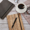 A5 wood look notebook in natural wood tones, a black notebook, a pen, coffee cup, glasses, and a magazine on a table.