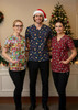 Three models wear festive unisex scrub tops in red, navy, and multicoloured designs, with a Christmas tree in the background.