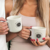 A coffee mug in light grey with a logo, held by two people. Featuring a round design and a comfortable handle.