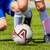 A soccer ball with a red and black design on a grassy field, near players in blue and white uniforms.