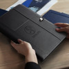 A black leather portfolio with a logo, held by hands, placed on a wooden table with blue folders nearby.