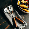 Stainless steel salad servers with wooden handles resting on a grey linen napkin, alongside a bowl of fresh fruit.