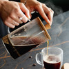 A coffee plunger with a wooden lid is being used to pour hot coffee into a clear glass mug.