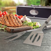 A stainless steel BBQ set with a spatula and fork, displayed beside grilled meat, tomatoes, and herbs. The set is in a black case.