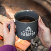 A matte enamel coffee mug in dark grey, held with two hands, filled with a dark beverage near a campfire.