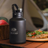 A drink bottle in matte black with a handle and silver cap, featuring a logo. Nearby are fruits, salad, and grilled food.