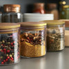 A set of large round storage canisters with transparent bodies and wooden lids, containing various spices and herbs.