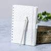 A white spiral-bound notebook with a textured cover, accompanied by a matching pen, set against a natural stone surface.