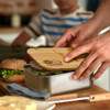 A stainless steel lunch box with a bamboo lid, filled with sandwiches and greens, on a wooden surface.