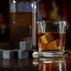 A glass with whiskey and grey whiskey stones beside a drink bottle and a stainless steel tongs.