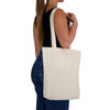 A cream tote bag held by a person, featuring shoulder straps and a simple design. The bag has a logo.