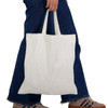 A white tote bag with short handles, held by a person wearing blue jeans and sneakers. The bag features a logo.