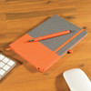 A grey notebook with an orange cover and an orange pen placed on a wooden desk beside a keyboard and mouse.