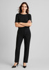 A woman wears black comfort wool stretch relaxed pants paired with a black top and high heels, standing confidently.
