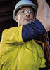 A man wearing a bright yellow long sleeve work shirt with navy blue accents, accompanied by safety glasses and a hard hat.
