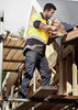 A man wearing a high-visibility yellow and black shirt and navy cargo pants uses a tool while standing on scaffolding.