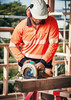 A worker in a high-visibility orange long-sleeve polo with reflective tape operates a circular saw on a construction site.