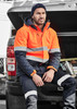 A man wearing an orange and navy unisex hi-vis softshell jacket sits on the back of a vehicle.