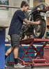 A man in navy shorts works on a motorcycle in a garage, featuring a logo on the shorts.