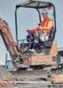 A worker in a high-visibility orange shirt and white hard hat operates a small excavator on a construction site.
