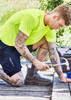 A vibrant yellow men's hi-vis tee worn by a tattooed man, kneeling and using a hammer on a wooden surface.