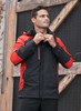 A man wearing a black and red jacket, standing against a rustic wooden backdrop. The jacket has a hood and zippers.
