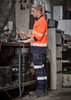 A woman wearing high-visibility orange and navy cargo pants with cuffs, working at a bench in a workshop.