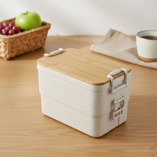 A beige bamboo stackable lunch box with a wooden lid, placed on a wooden surface beside a woven basket and cup.