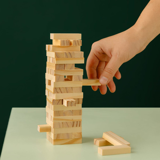 A hand removes a wooden block from a tall, stacked tower of light-coloured Jenga pieces on a green background.