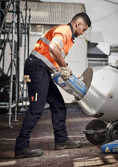 A man in orange and grey high-visibility workwear pours from a bag into a mixer, wearing navy rugged cargo pants.