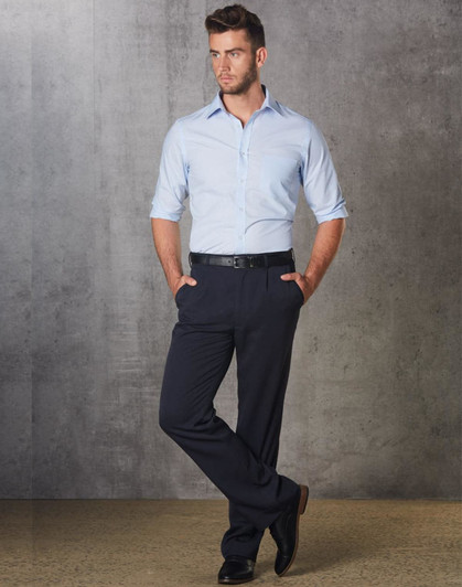 A man in a light blue shirt and black permanent press pants stands with hands in pockets against a grey backdrop.