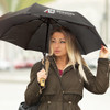 A black compact umbrella held by a woman in a brown coat, featuring a logo on the canopy.