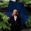A navy umbrella with a unique design, held by a person in a black blazer, surrounded by lush greenery.