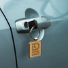 A square key ring with a metallic finish, attached to a car door handle. Features a branding logo.