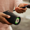 A Bluetooth speaker with a black exterior and a vibrant green light, held in one hand while a smartphone is in the other.
