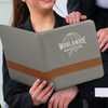 A grey portfolio with a brown strap, held by a person, featuring a logo on the front.