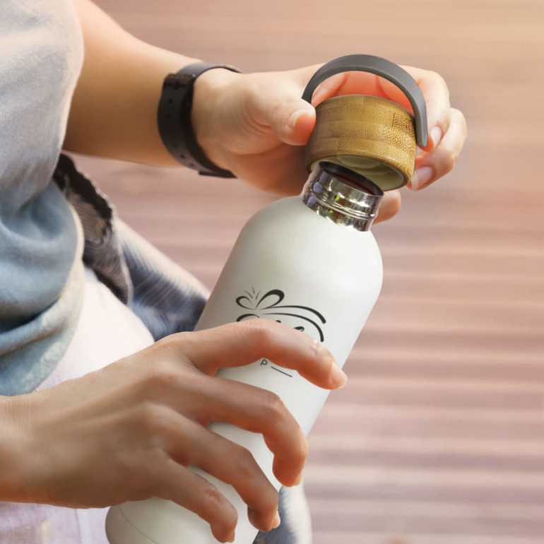 A drink bottle made of stainless steel, featuring a white body and a bamboo lid, held by a person.