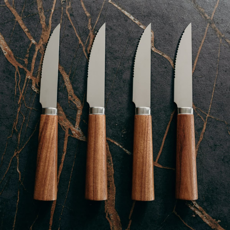A steak knife set featuring four stainless steel knives with brown wooden handles, arranged on a dark stone surface.