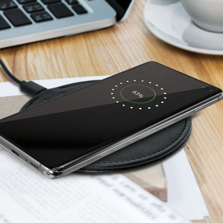 A black wireless charger with a smartphone on top, displaying a charging indicator, on a wooden desk with a laptop and cup.