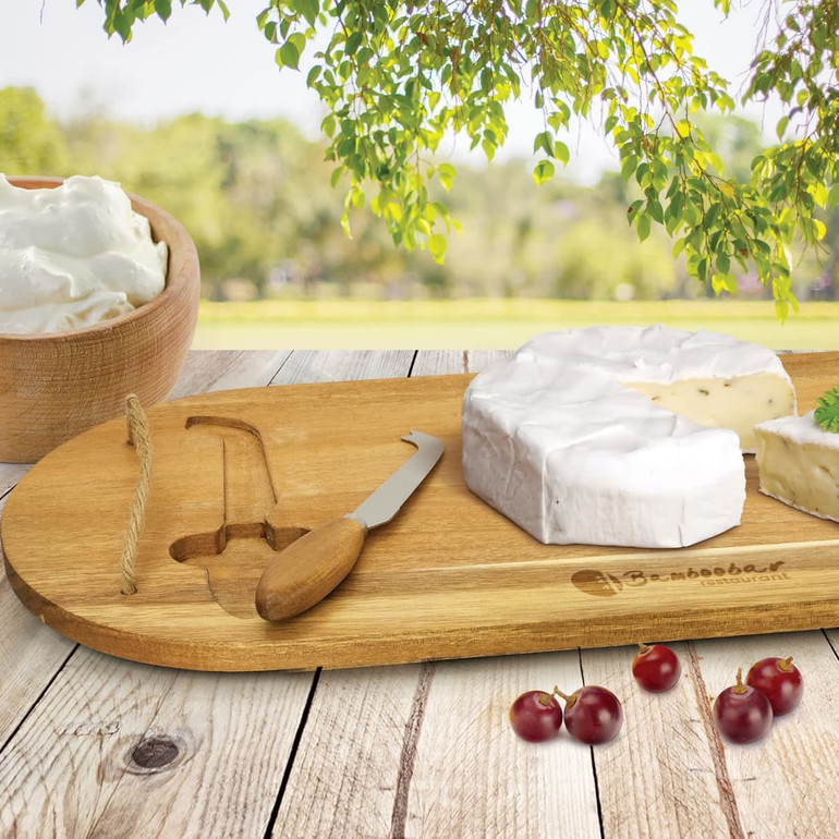 A wooden cheese board with a knife, featuring a round cheese and grapes. The board has a logo and is set outdoors.