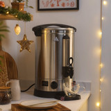 Stainless steel hot water urn with tap, on kitchen counter near glass cup, festive decor