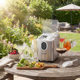 Countertop ice maker in stainless steel on garden table with summer drinks and fruit.