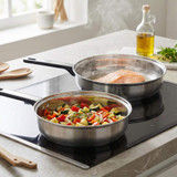 Stainless steel frying pans on induction hob, cooking salmon fillet and mixed vegetables.